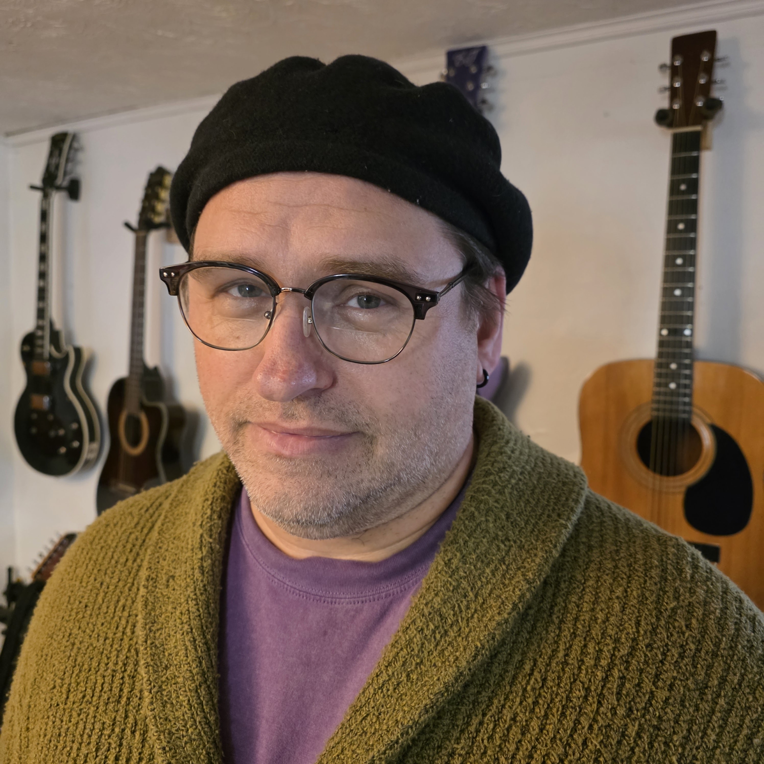 John_Shaver Photo of Emery- A short haired person in 70s swirls and tatoos talks into a microphone while in their wheelchair. They look and are super important. The lighting is as colorful as they are.