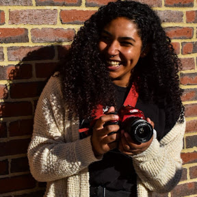 A photo of Avery, sitting on the ground leaned against a neutral wall. She is a white woman with shoulder lenght brown hair and a quirky smile. 